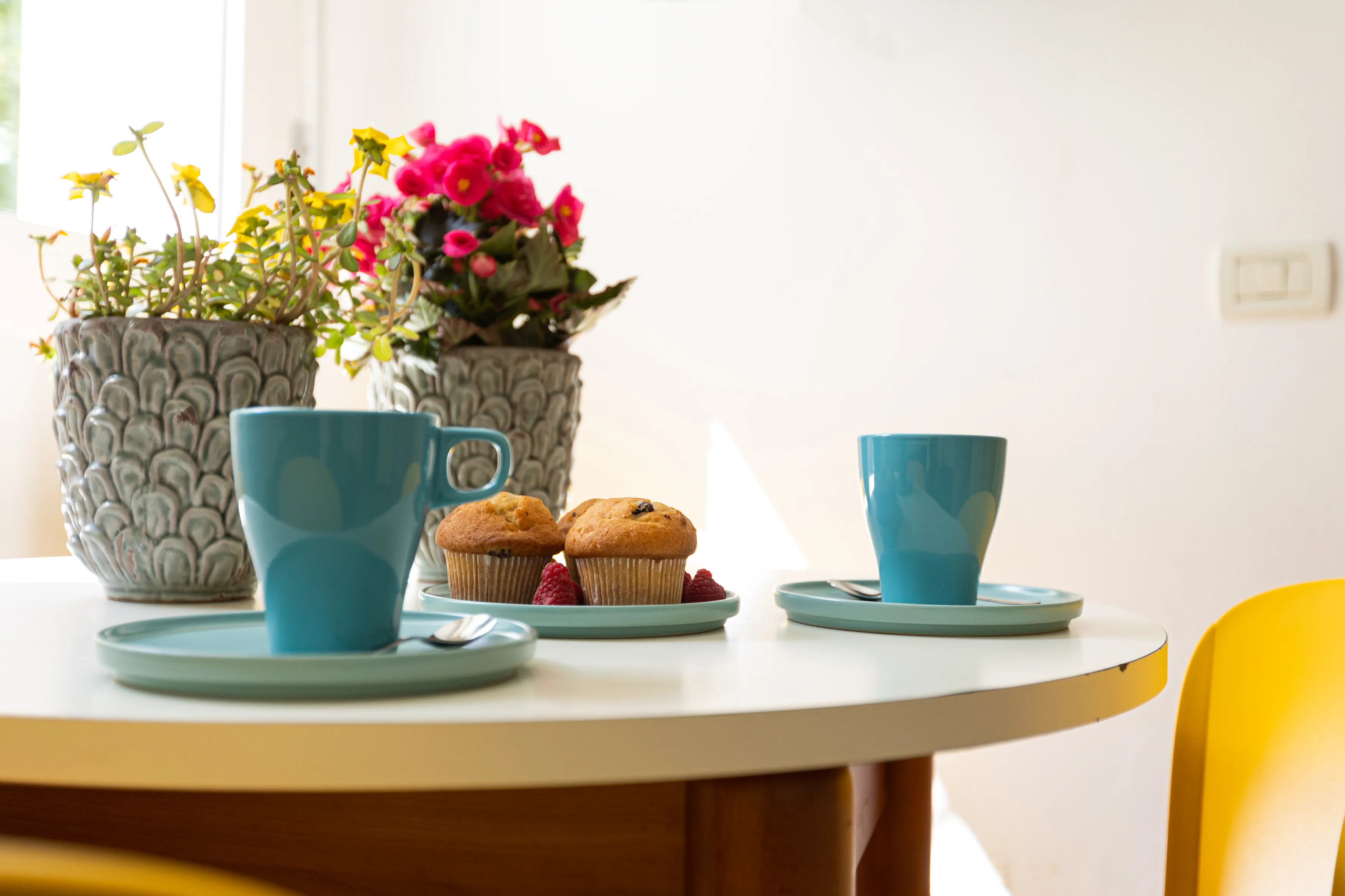 Tavolo bianco con due tazze azzurre su piattini con cucchiaini, due muffin con lamponi e due vasi di fiori gialli e rosa sullo sfondo.