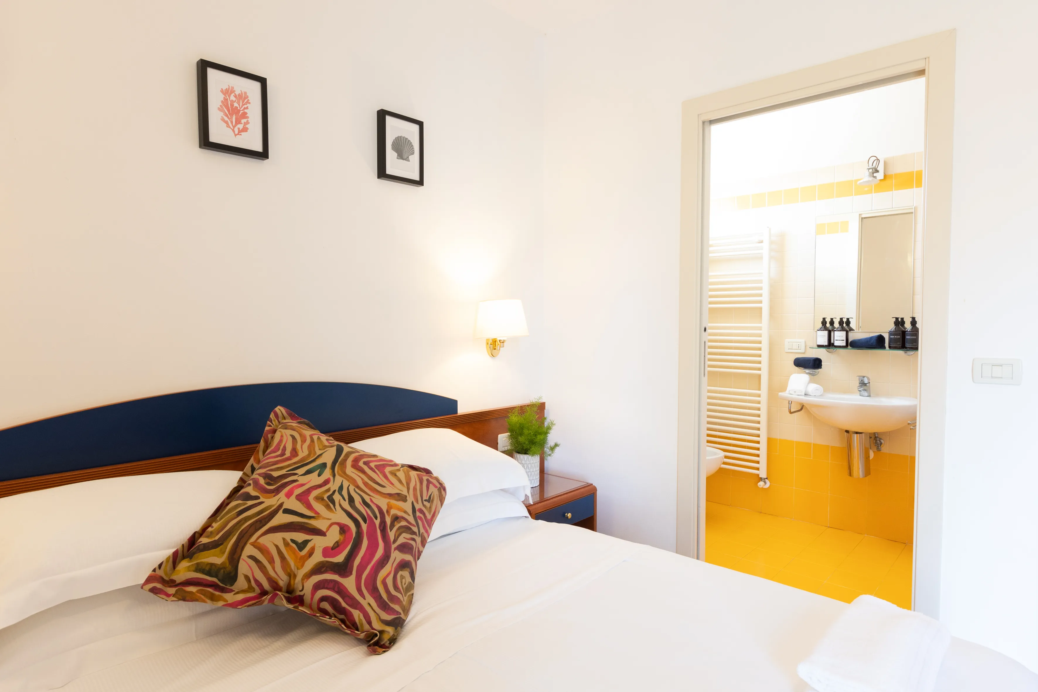 Camera con letto matrimoniale, armadio blu, frigorifero bianco e finestra luminosa su parete bianca.