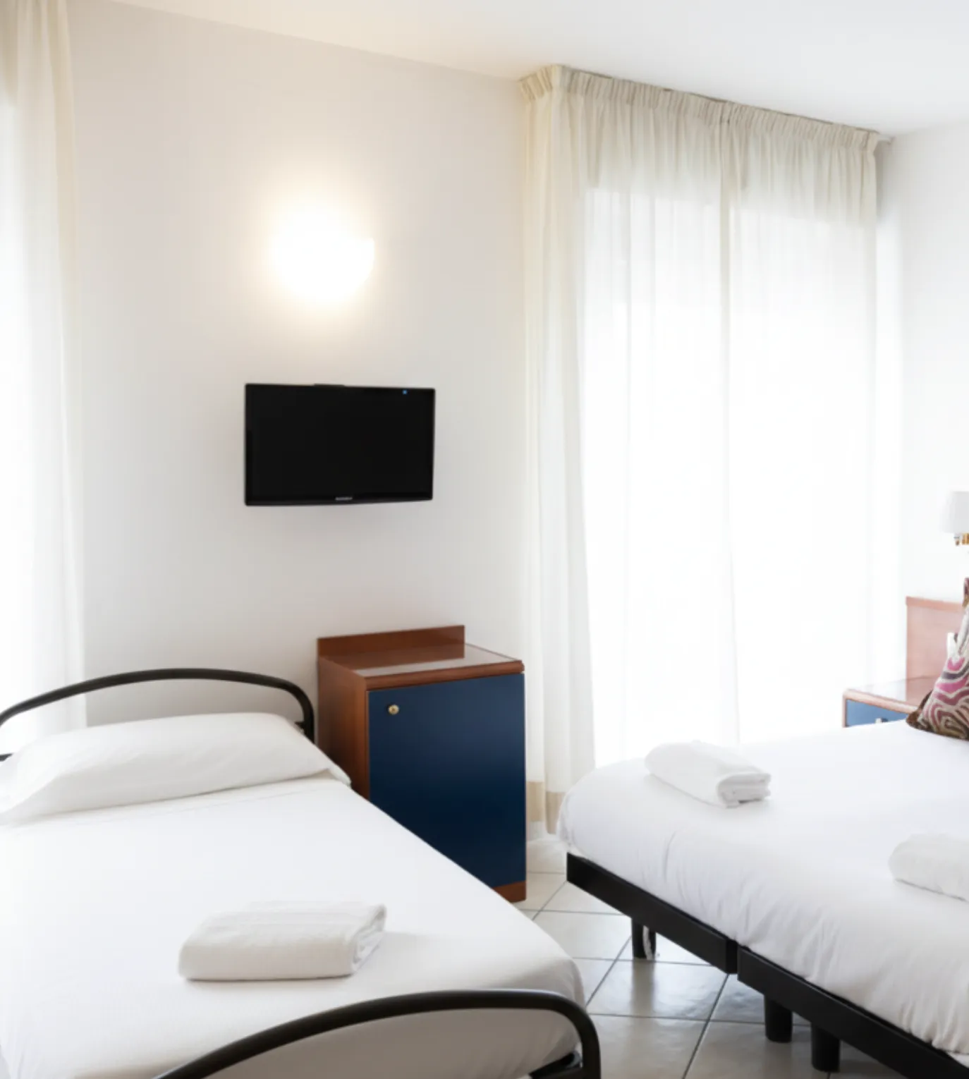 Camera luminosa con due letti singoli, lenzuola bianche, asciugamani piegati e una TV a schermo piatto montata su una parete bianca.