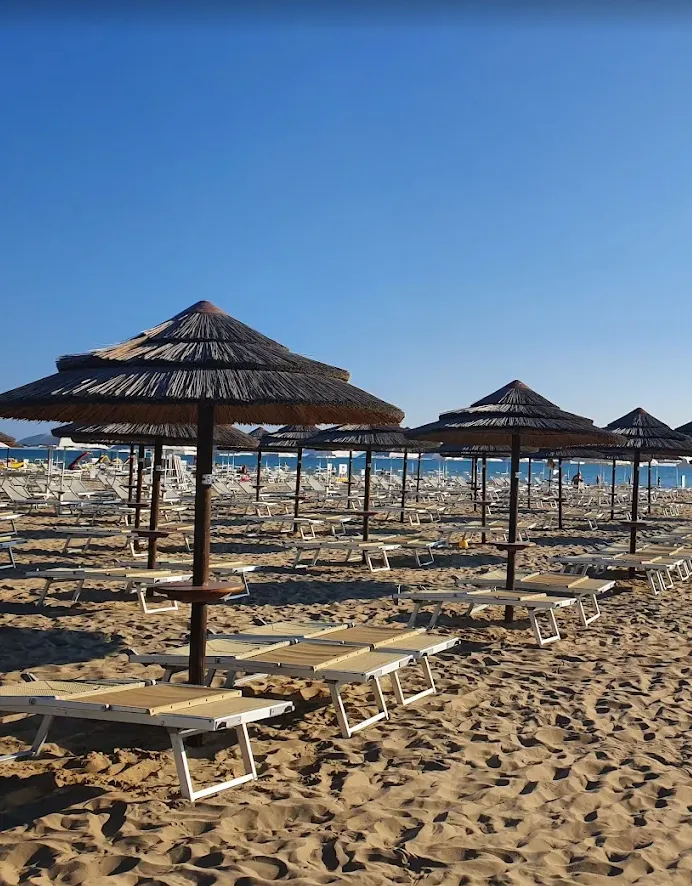 Ombrelloni di paglia e lettini disposti ordinatamente su una spiaggia sabbiosa con il mare e il cielo azzurro sullo sfondo.