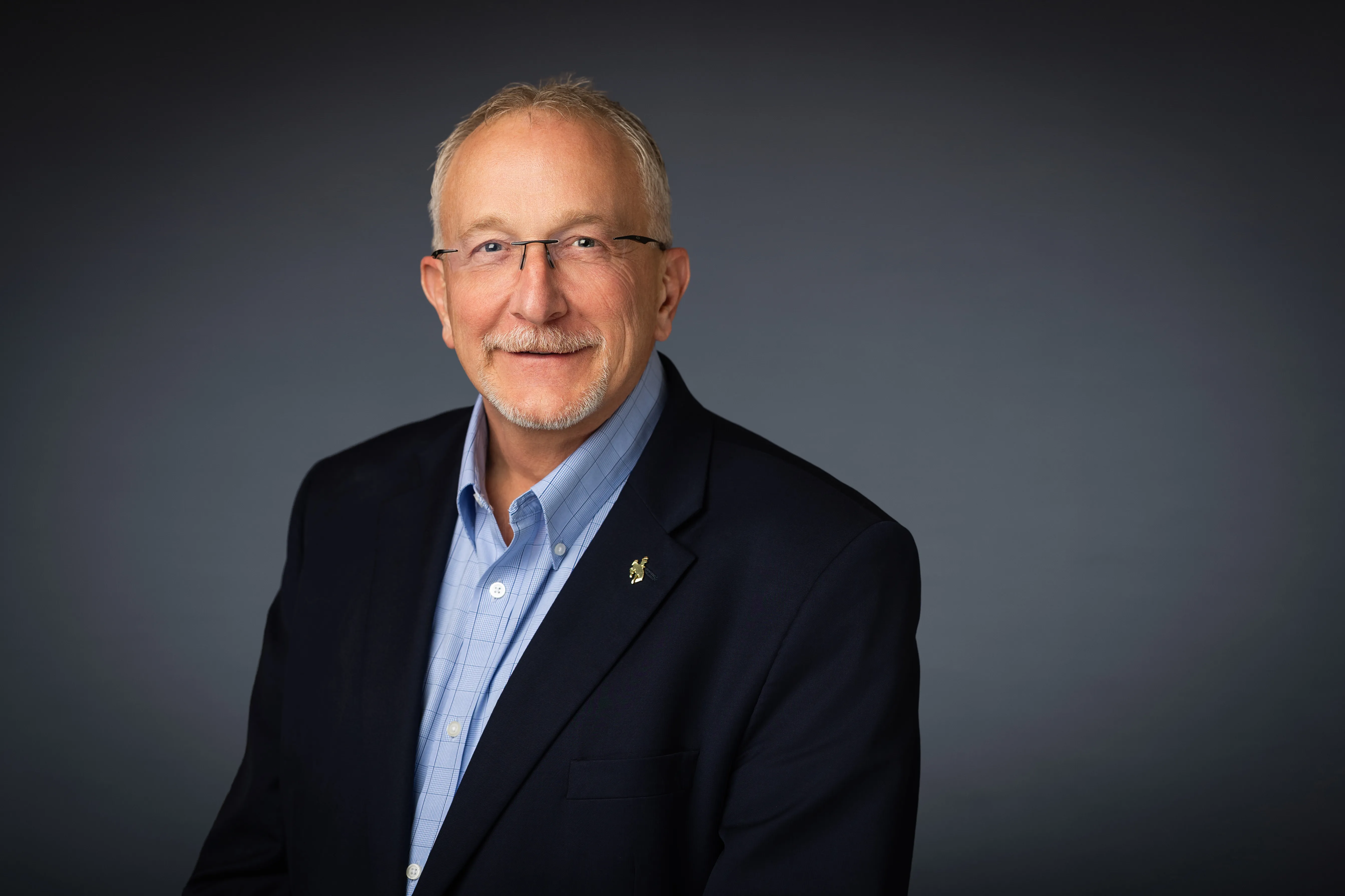 Smiling middle-aged man with glasses, short gray hair, and a beard wearing a navy blazer and light blue checkered shirt against a gray background.