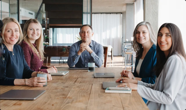 Five businesspeople smiling and sitting at a wooden conference table in a modern office.