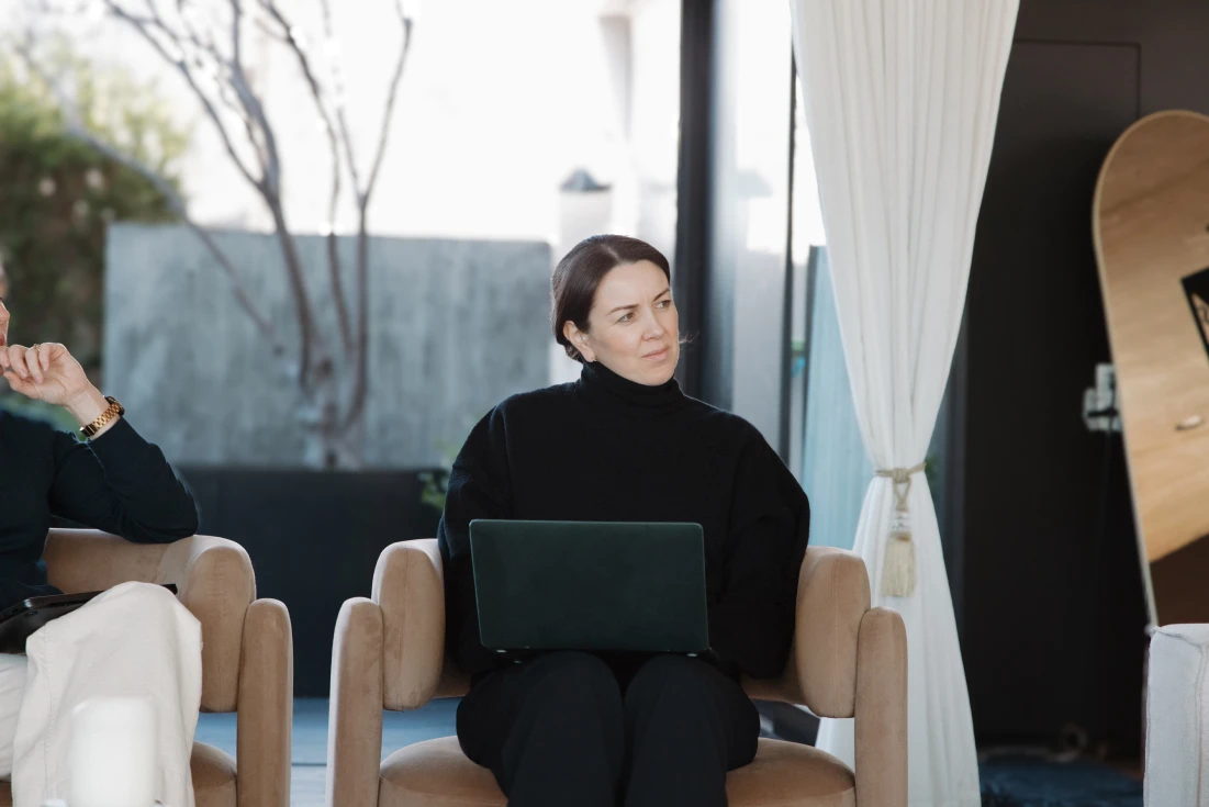 Woman in a black turtleneck sitting on a beige chair with a laptop on her lap, looking thoughtfully to the side.