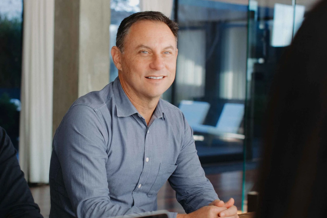 Smiling man in a blue button-up shirt sitting at a table with hands clasped indoors.