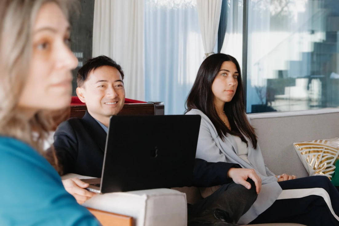 Three people sitting on couches engaged in a conversation, with one man using a laptop.
