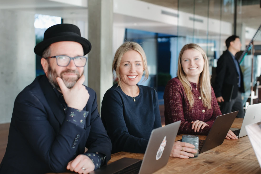Three smiling people sitting at a wooden table with laptops in a modern office setting, one man wearing a black hat and glasses, two women holding a glass and resting hands on laptops.