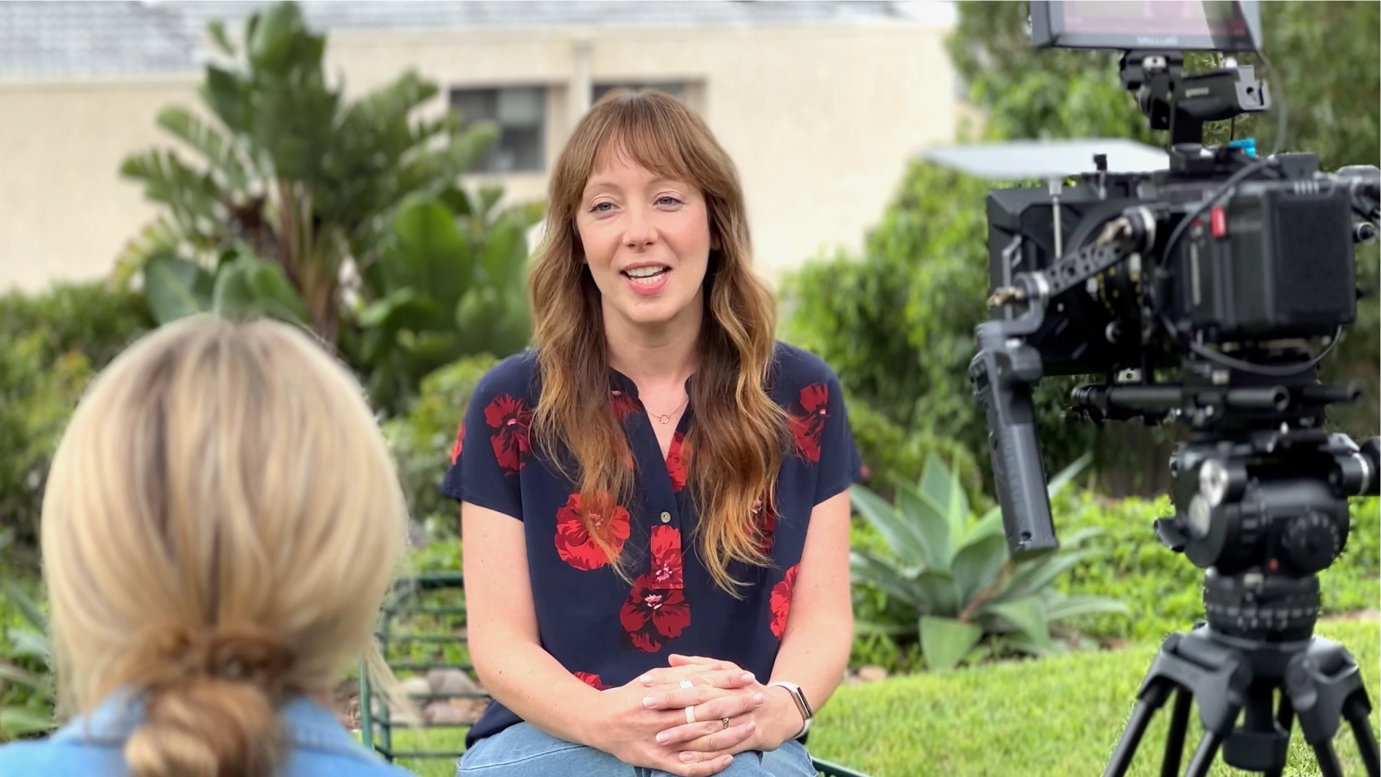 Woman with long brown hair and a navy blouse with red flowers being filmed outdoors, speaking to another woman.