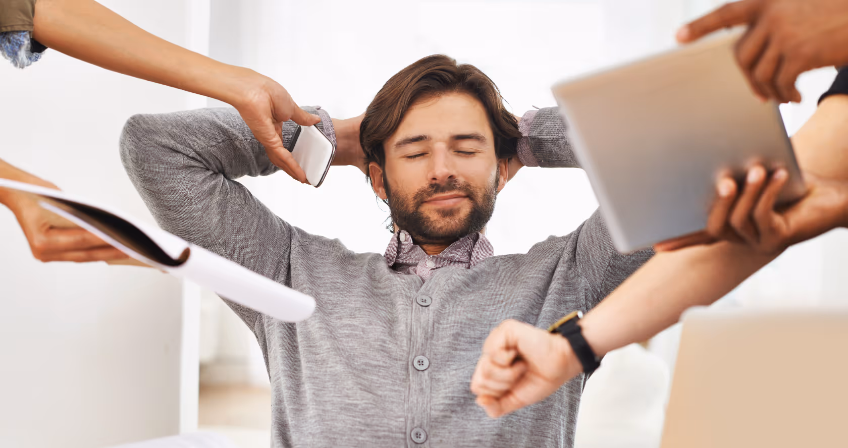 Man in a gray sweater leaning back with eyes closed while multiple hands around him hold a phone, documents, a tablet, and check a wristwatch.