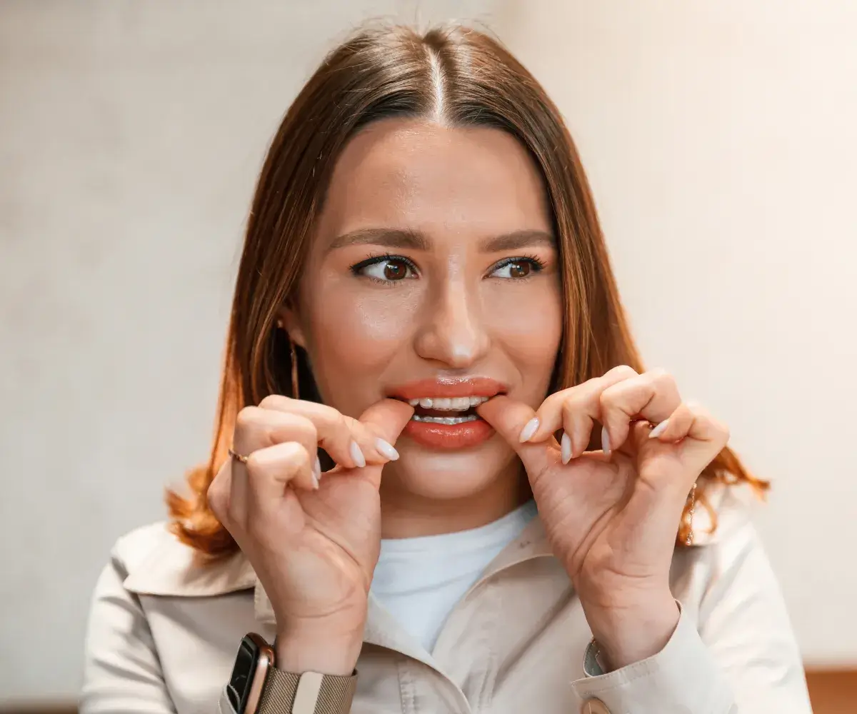 A woman biting her nails, wearing a light jacket and a watch, indoors.