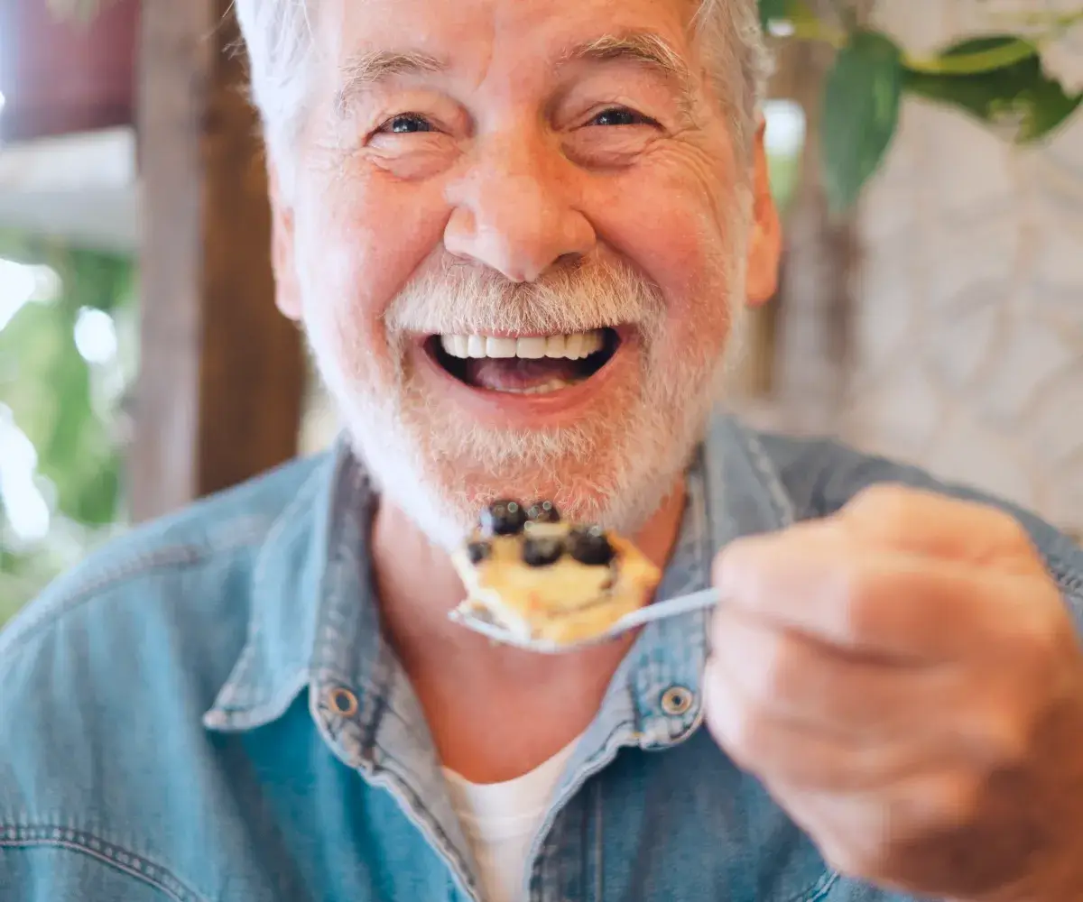 Smiling man holding a spoon with blueberry-topped dessert, wearing a denim shirt.