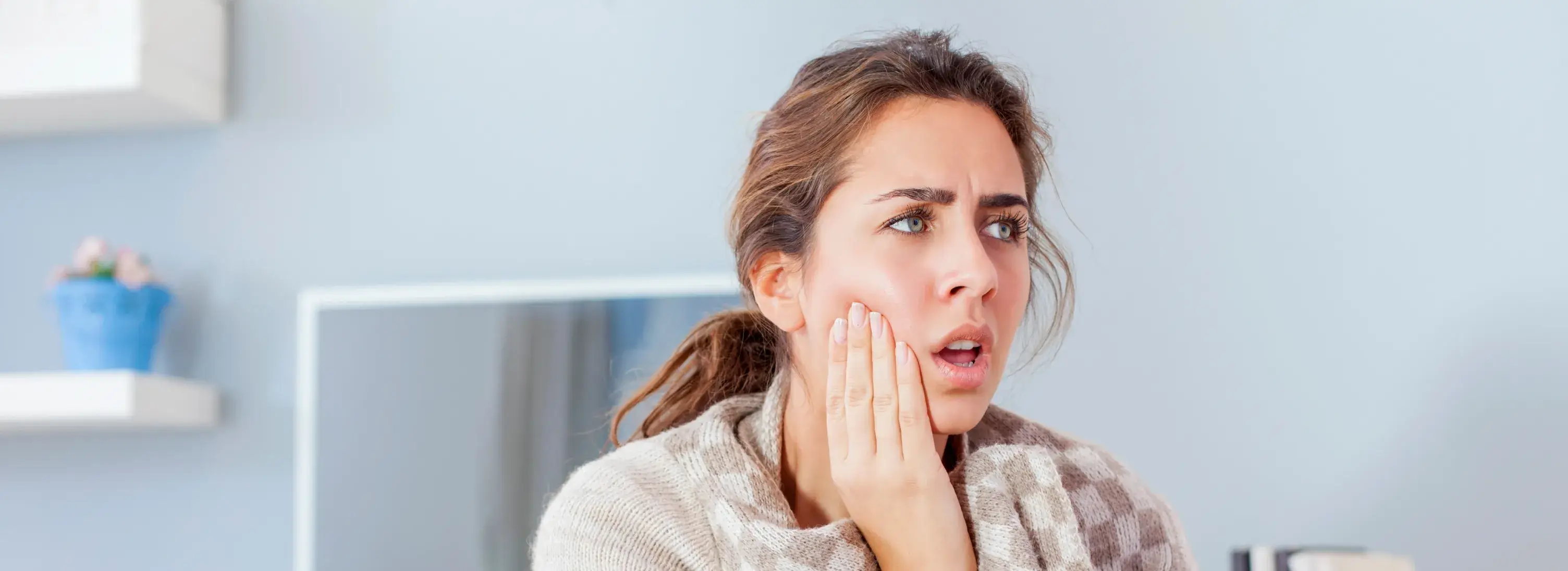 A woman sits, holding her cheek with a worried expression, possibly experiencing pain.