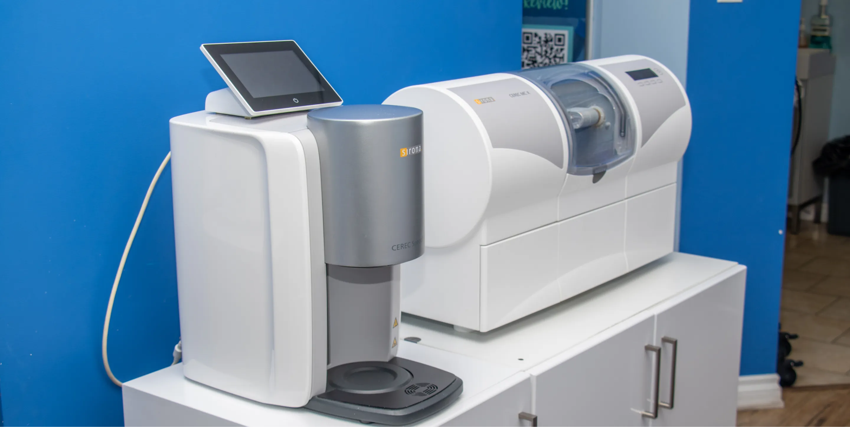 Dental milling machine and scanner on a white cabinet against a blue wall.