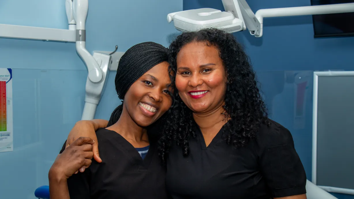 Two women wearing black scrubs smile and embrace in a dental office.
