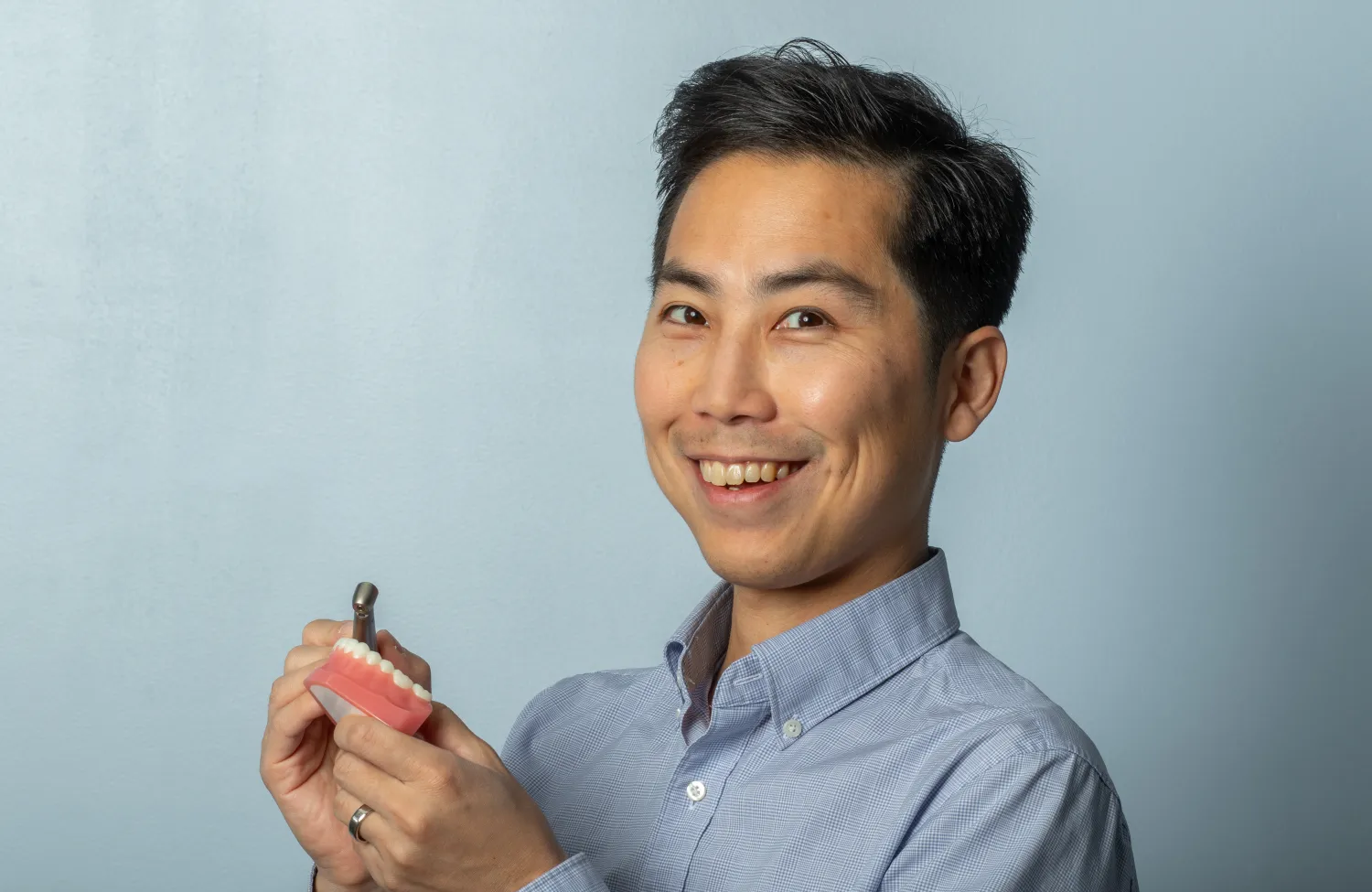 A person smiling while holding a dental model and toothbrush against a plain background.