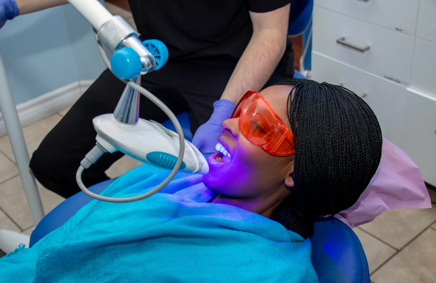 A person sits in a dental chair undergoing a teeth whitening procedure with protective eyewear.