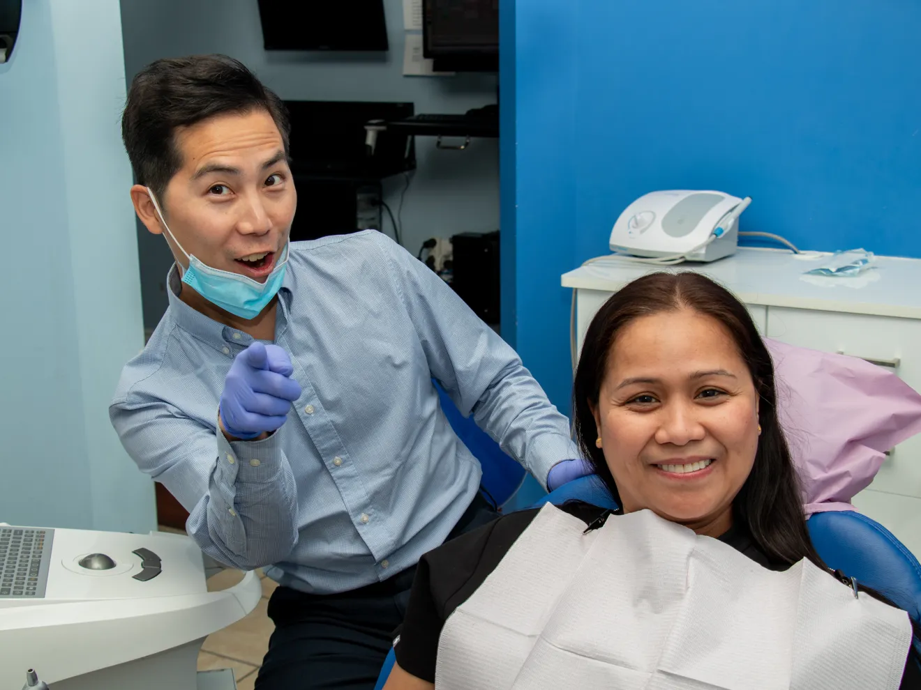 Smiling person holds mirror while sitting in a dental chair, with a dentist pointing at something.
