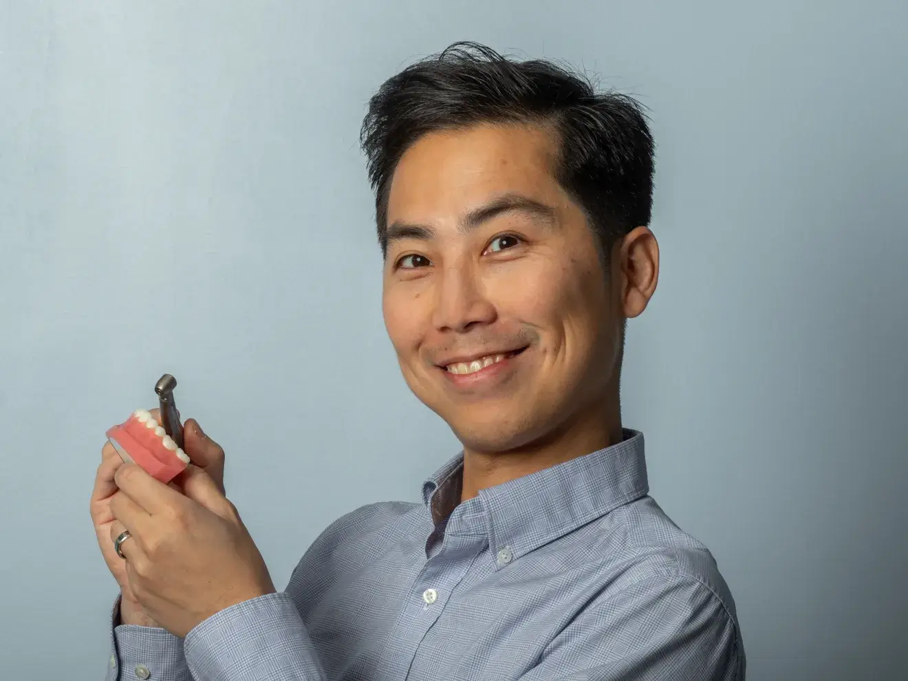 A smiling man holds a dental model and tool against a plain background.