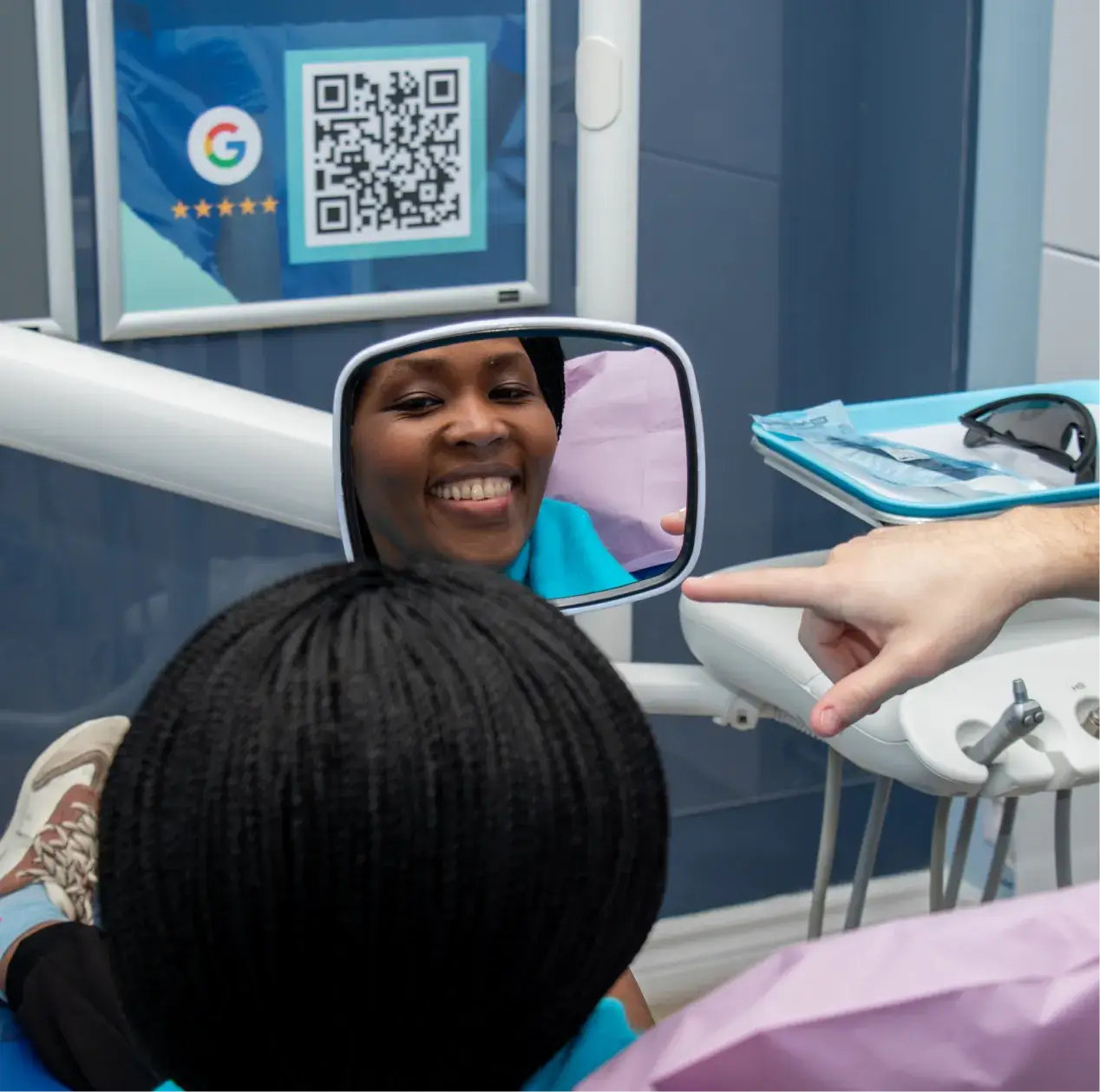 Person sitting in a dental chair, smiling and holding a mirror while a dentist points to it.