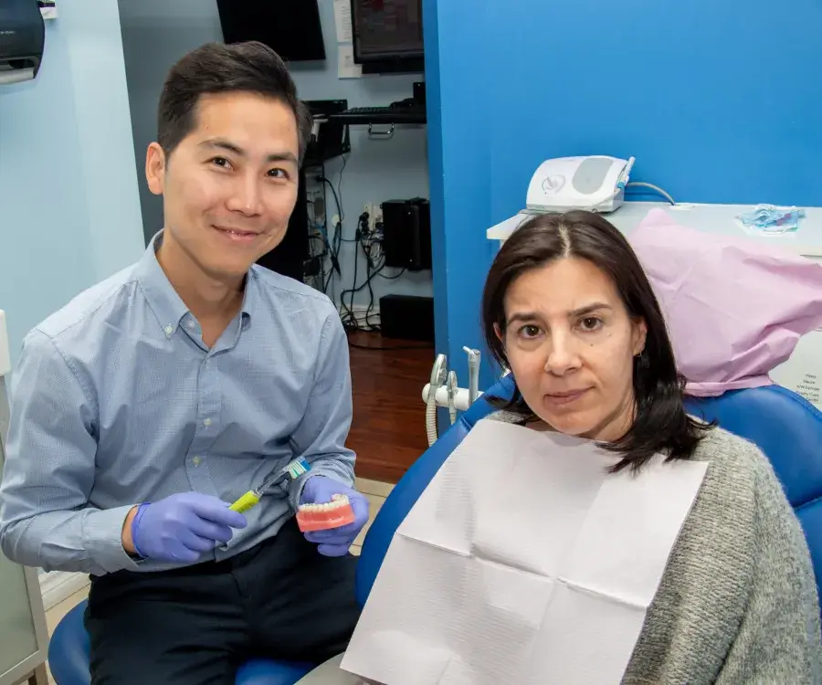 A dentist is holding dental tools and smiling next to a seated patient in a clinic.