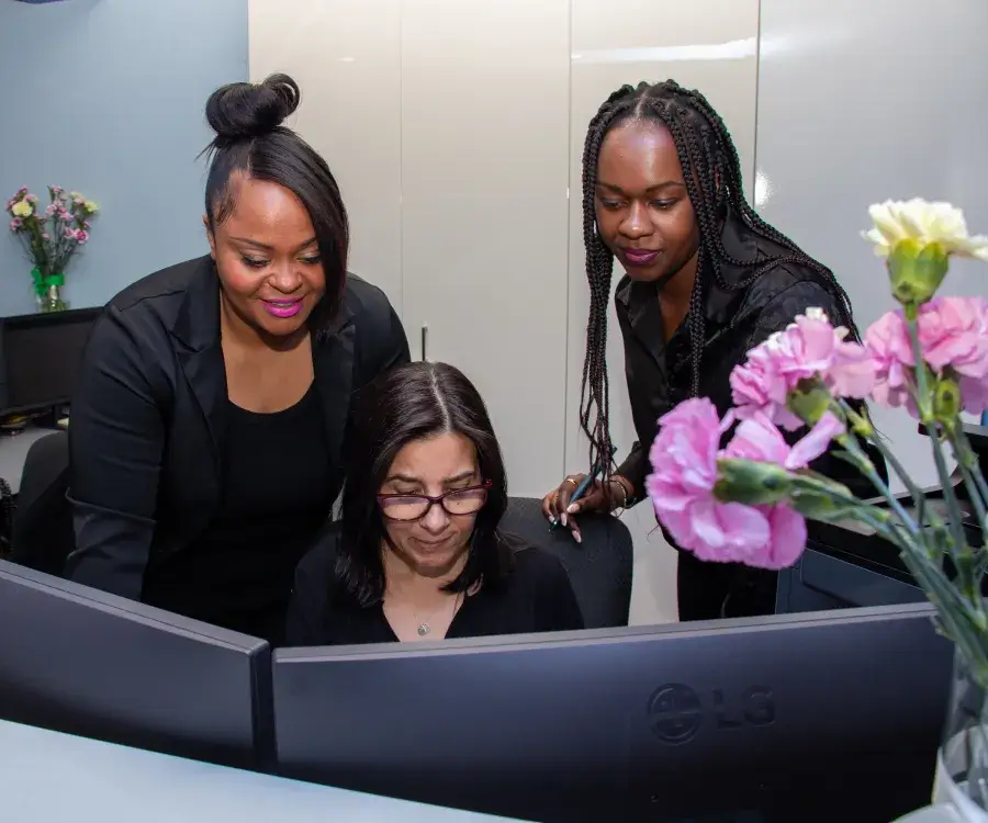 Three women in an office setting working together at a computer, with flowers on the desk.