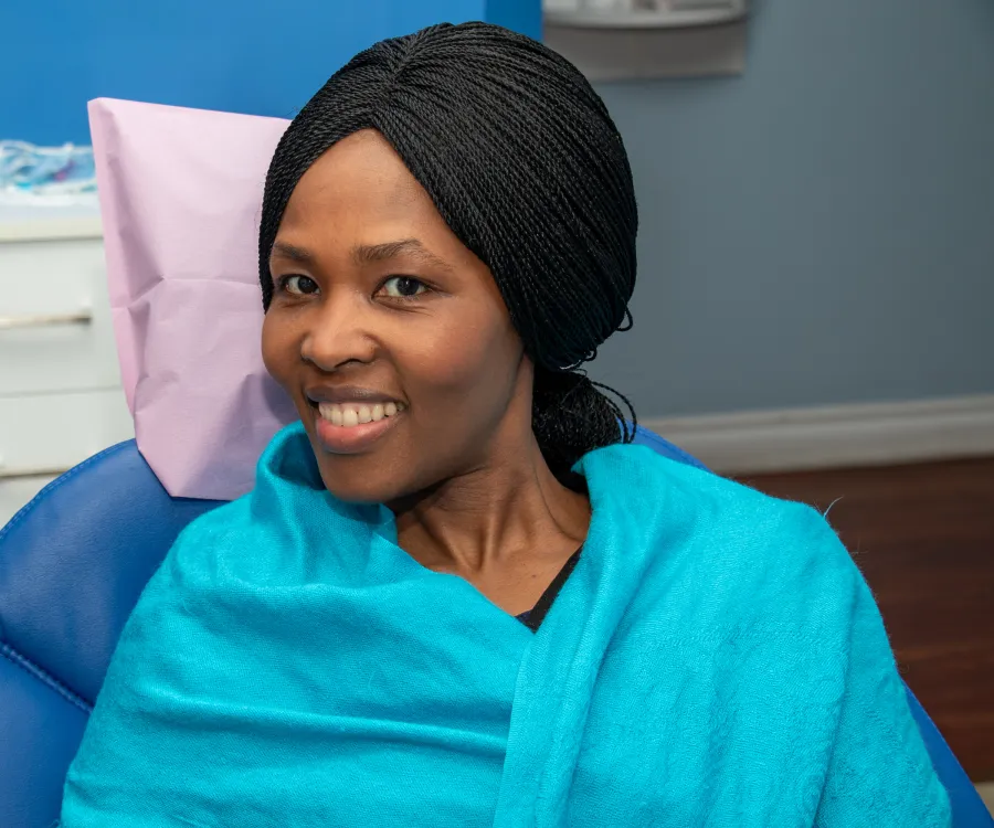 Smiling woman with braided hair sitting in a dental chair covered with a turquoise blanket.
