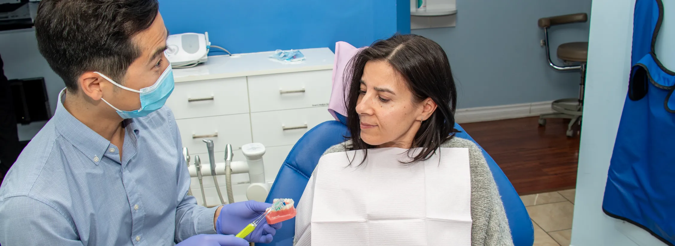 A dentist wearing a mask shows a dental model to a patient seated in a clinic chair.