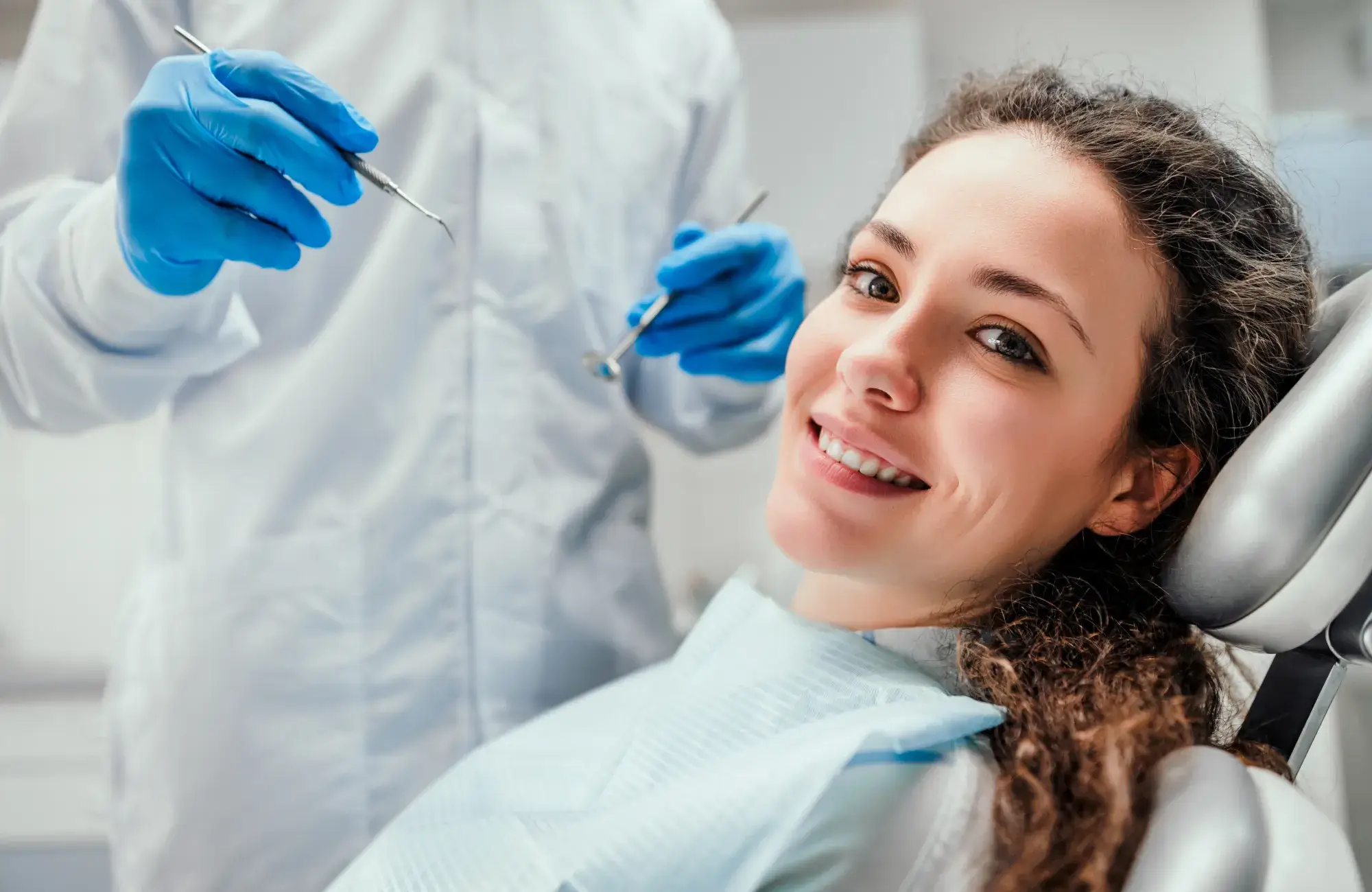 Woman sitting in a dentist's chair, smiling, while a dentist holds dental tools nearby.