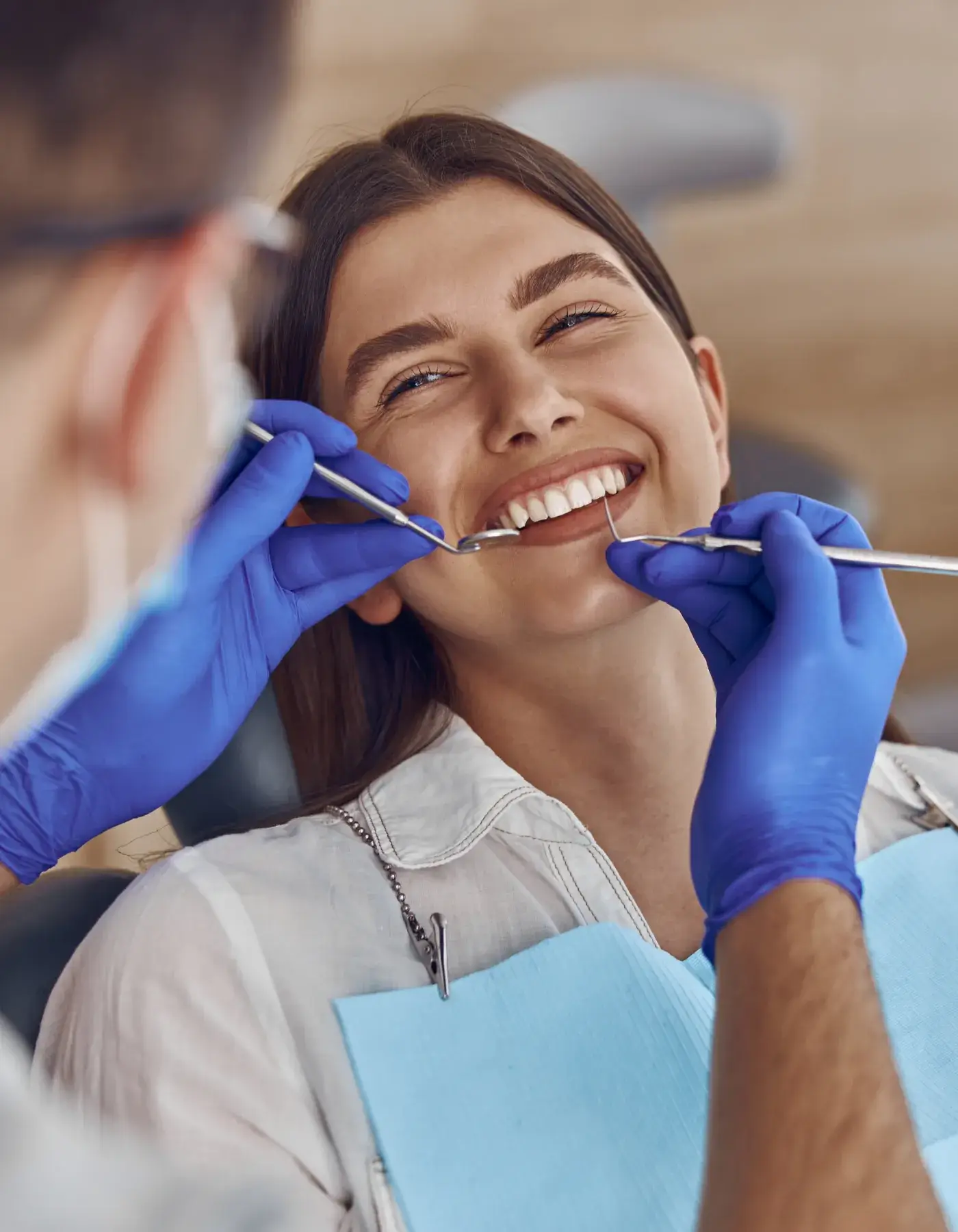 A dentist wearing gloves examines a smiling woman's teeth with dental tools.