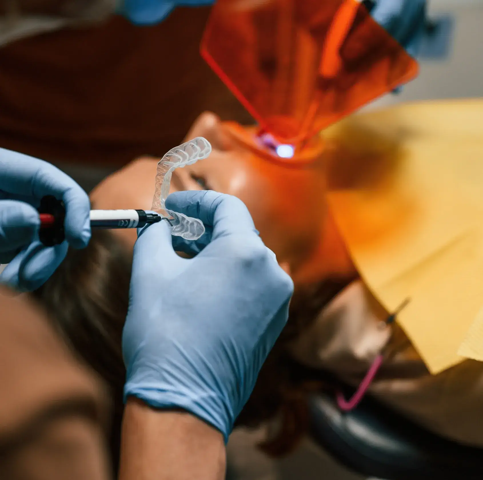 Dentist wearing gloves holds a dental guard and light curing tool near patient.