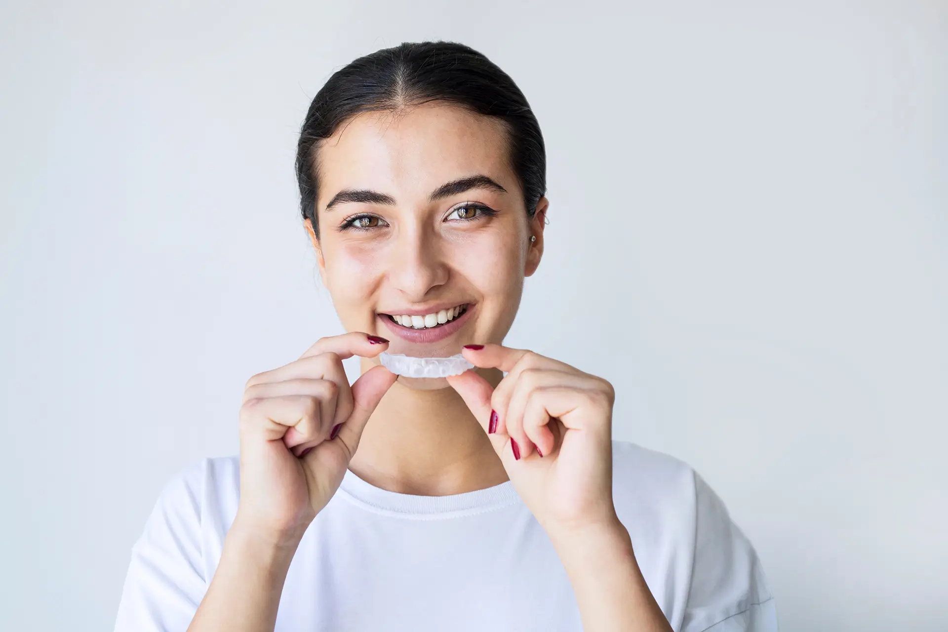 Person smiling while holding a clear dental aligner against a plain background.