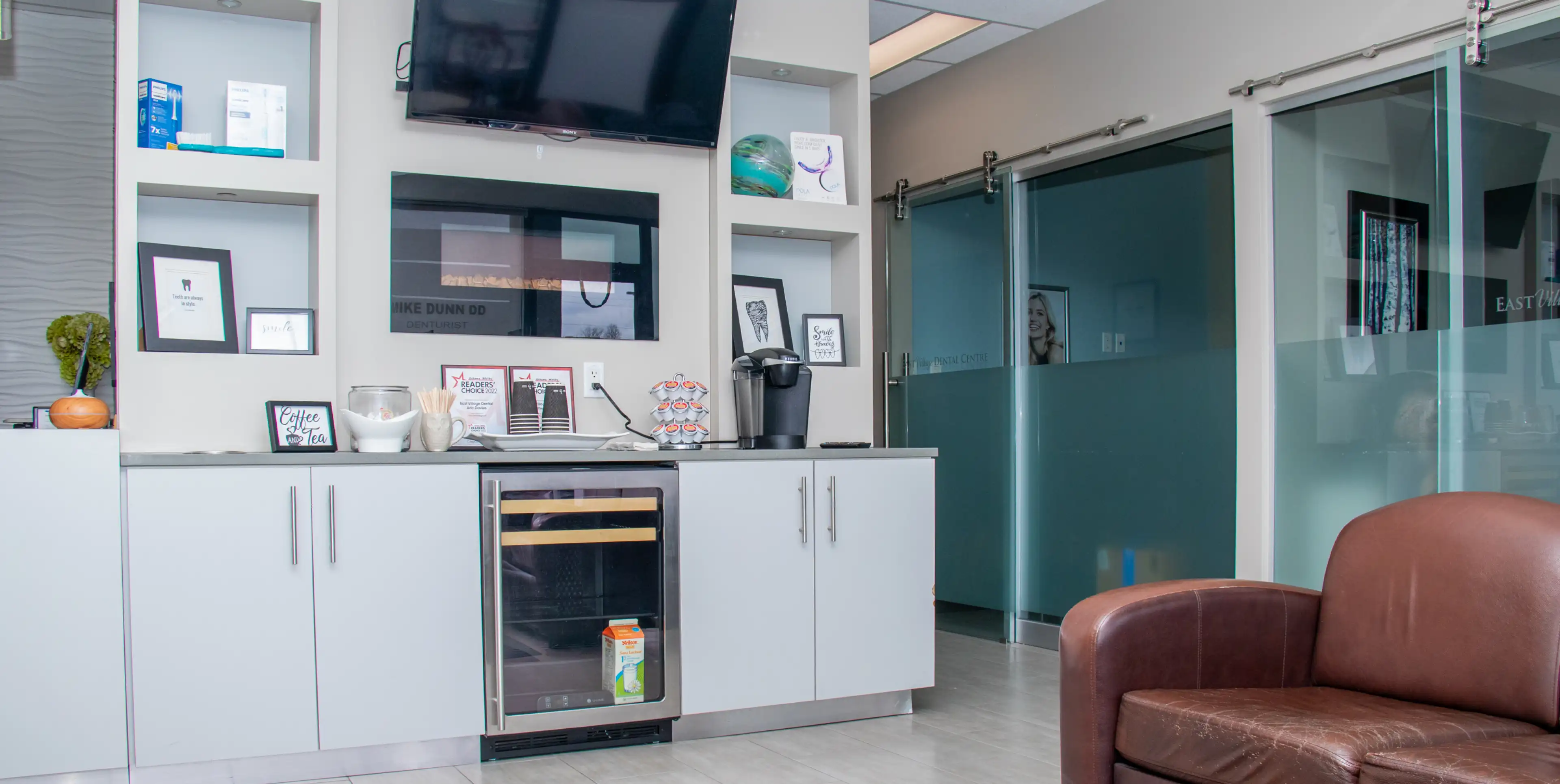 A waiting area with a coffee maker and a TV mounted above white cabinets.