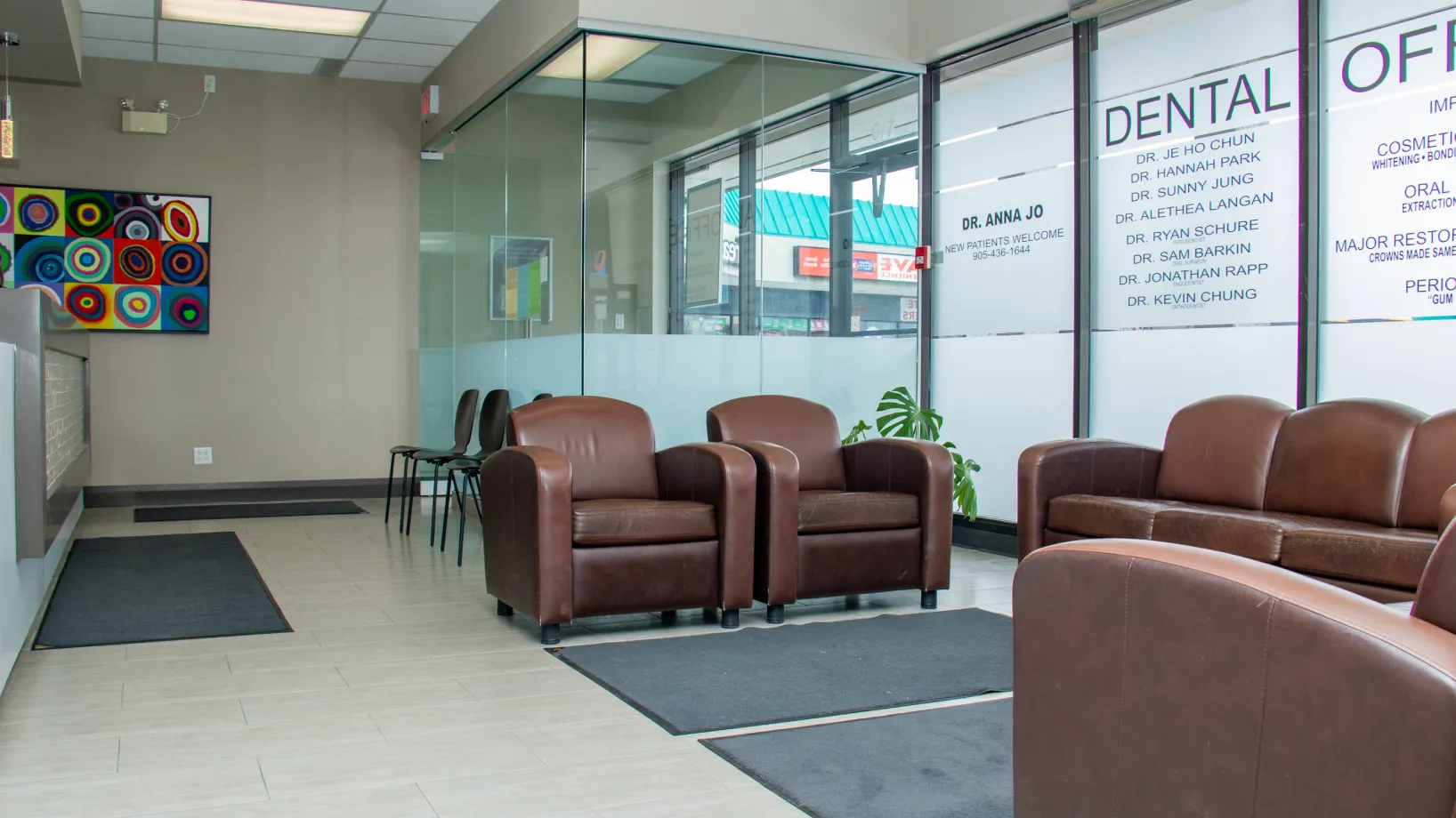 A dental office waiting area with brown chairs and a plant by a glass partition.