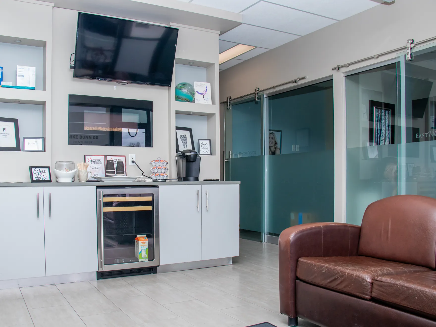 A waiting room with a TV, coffee machine, and leather chair. Glass doors lead to another room.