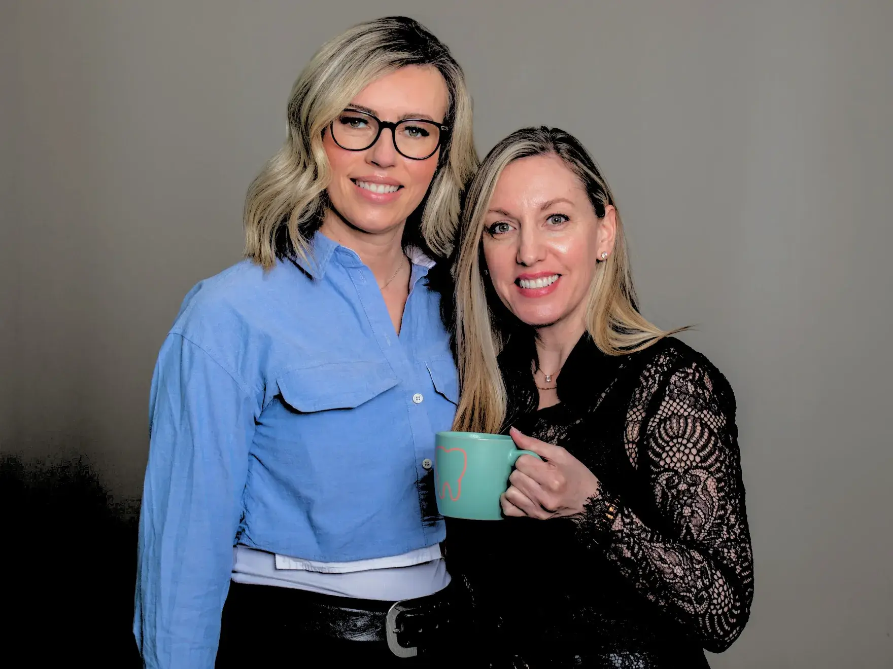 Two women smiling, one holding a teal mug, standing close together indoors.