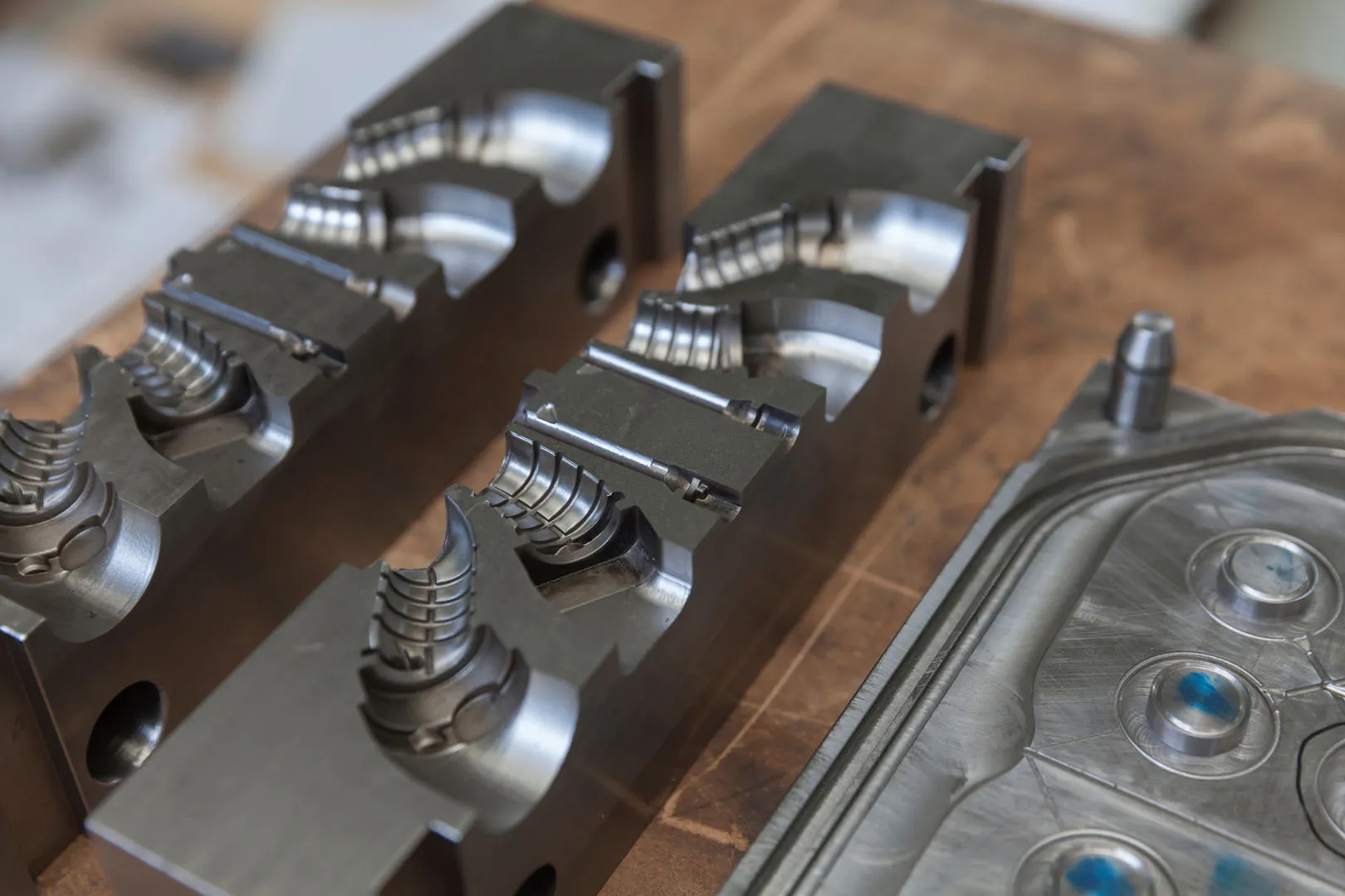 Close-up of two metal injection mold halves with detailed grooves and one mold plate on a wooden surface.