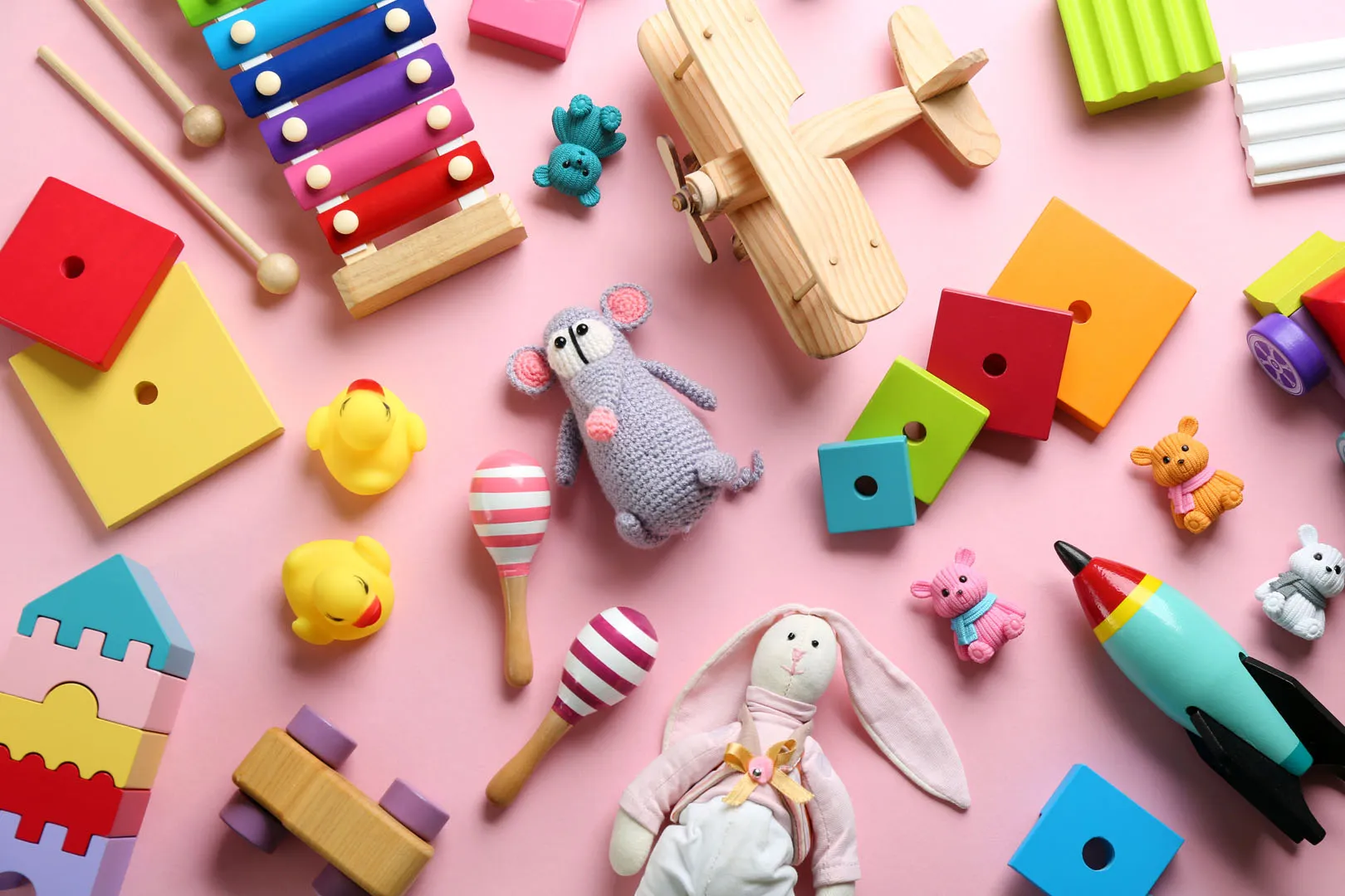 Assortment of colorful children's toys on a pink surface including a wooden airplane, xylophone, stacking blocks, rubber ducks, maracas, a toy rocket, and various stuffed animals.