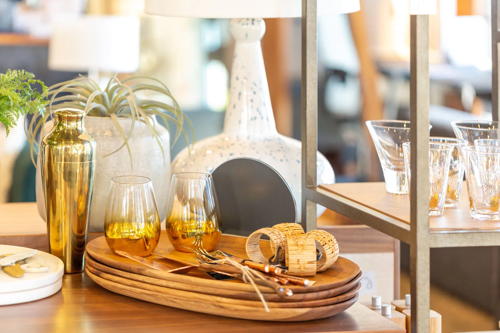 Wooden serving trays with two yellow-tinted glasses, cutlery with woven napkin rings, and a gold cocktail shaker on a wooden table with potted plants and glassware in the background.