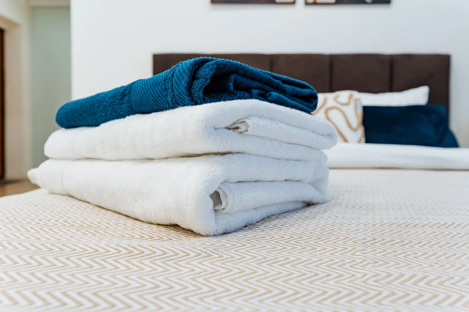 Stack of towels with two white towels and one folded blue towel on a beige patterned bedspread.