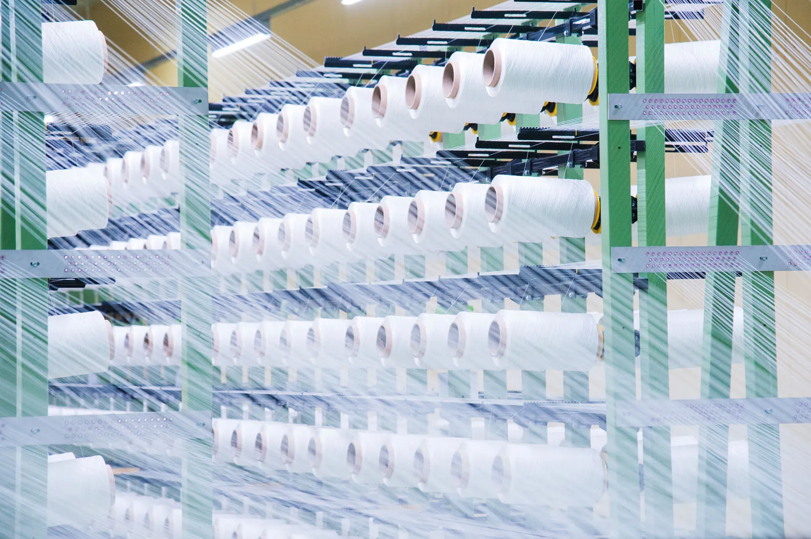 Rows of white thread spools on green racks with threads stretched across in a textile manufacturing setting.
