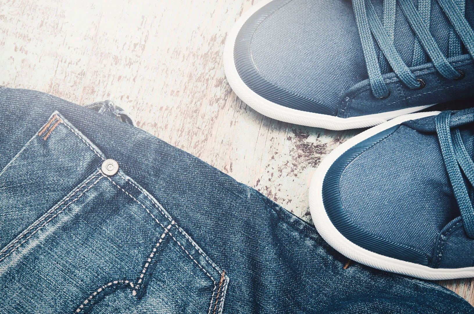 Close-up of blue denim jeans next to blue canvas sneakers on a wooden floor.