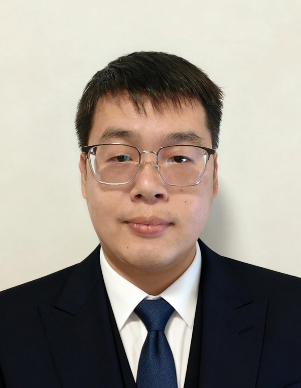 Portrait of a man with short black hair and glasses wearing a navy blue suit, white shirt, and dark blue tie against a plain light background.