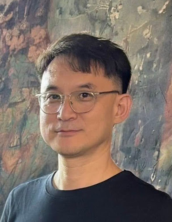 Portrait of a man with glasses and short black hair wearing a black shirt in front of a textured stone wall.