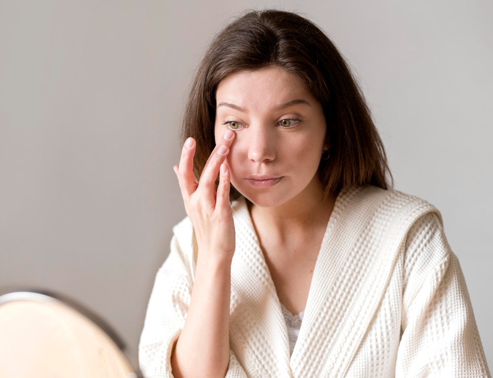 Woman in white waffle robe examining her face area in a mirror