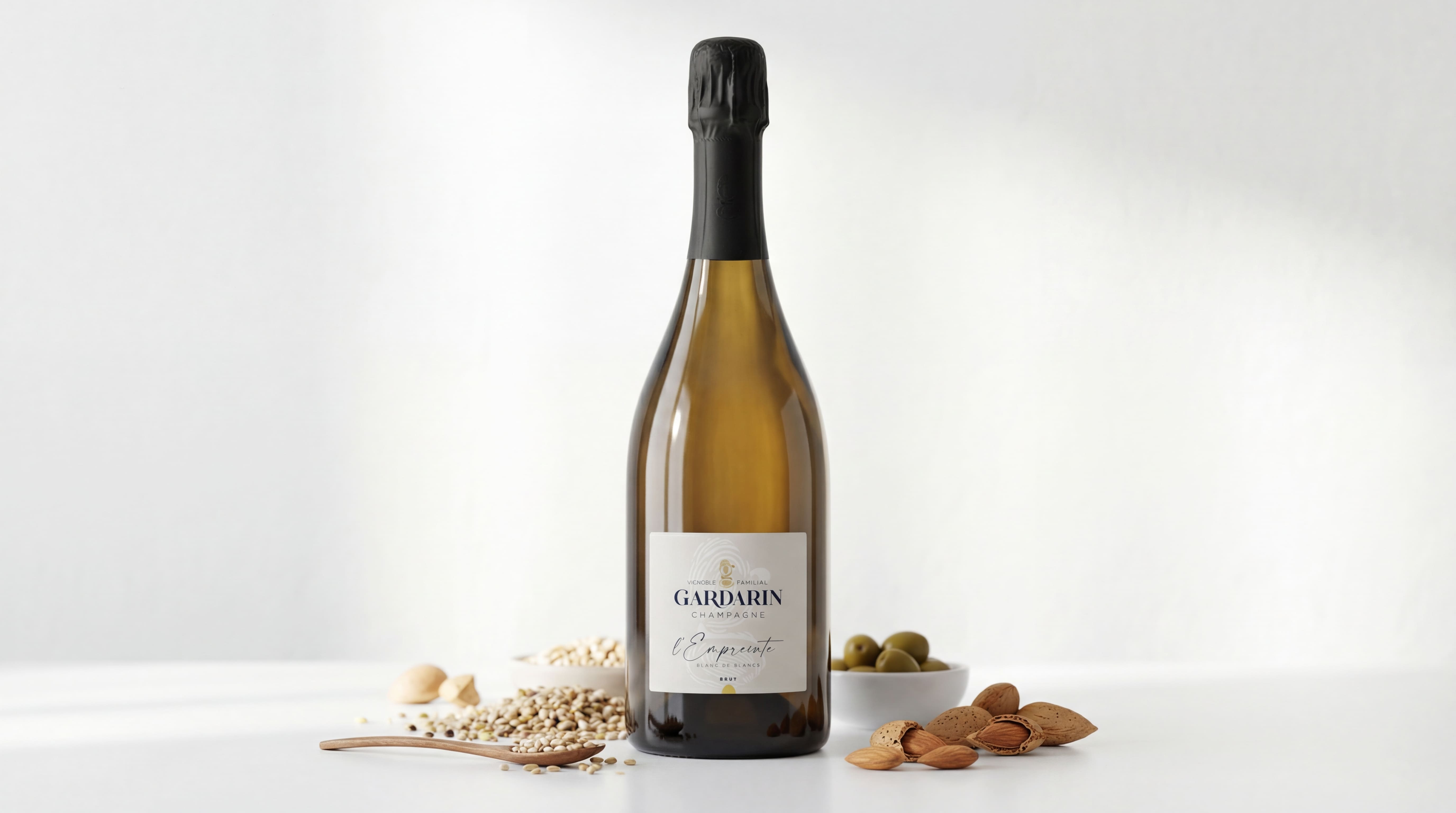 Bottle of Gardarin Champagne l'Empreinte surrounded by almonds, grains in a wooden spoon, and green olives in small white bowls on a white surface.