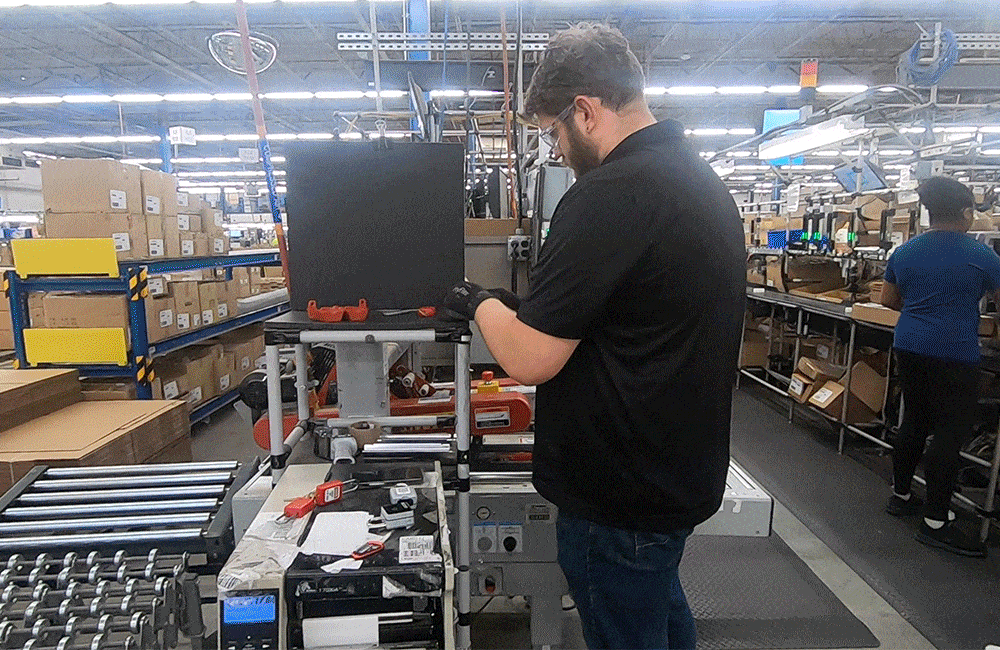 Man wearing gloves assembling or inspecting black product in a factory with conveyor belts and shelves.