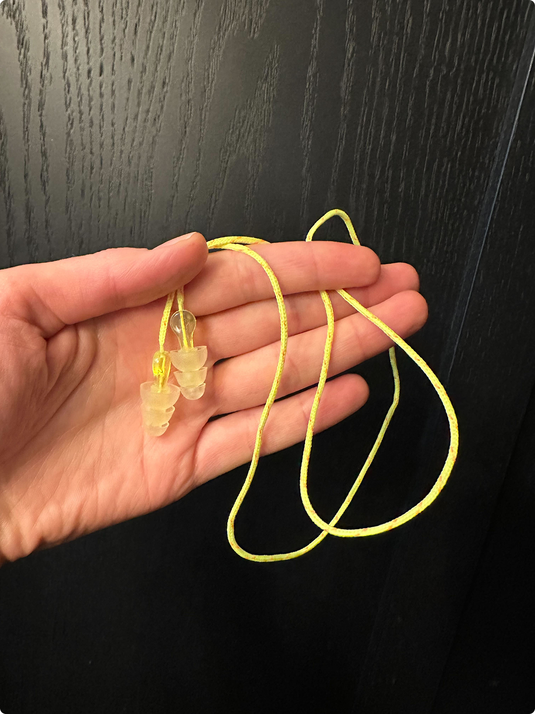Hand holding yellow corded reusable earplugs with translucent silicone tips against a black background.