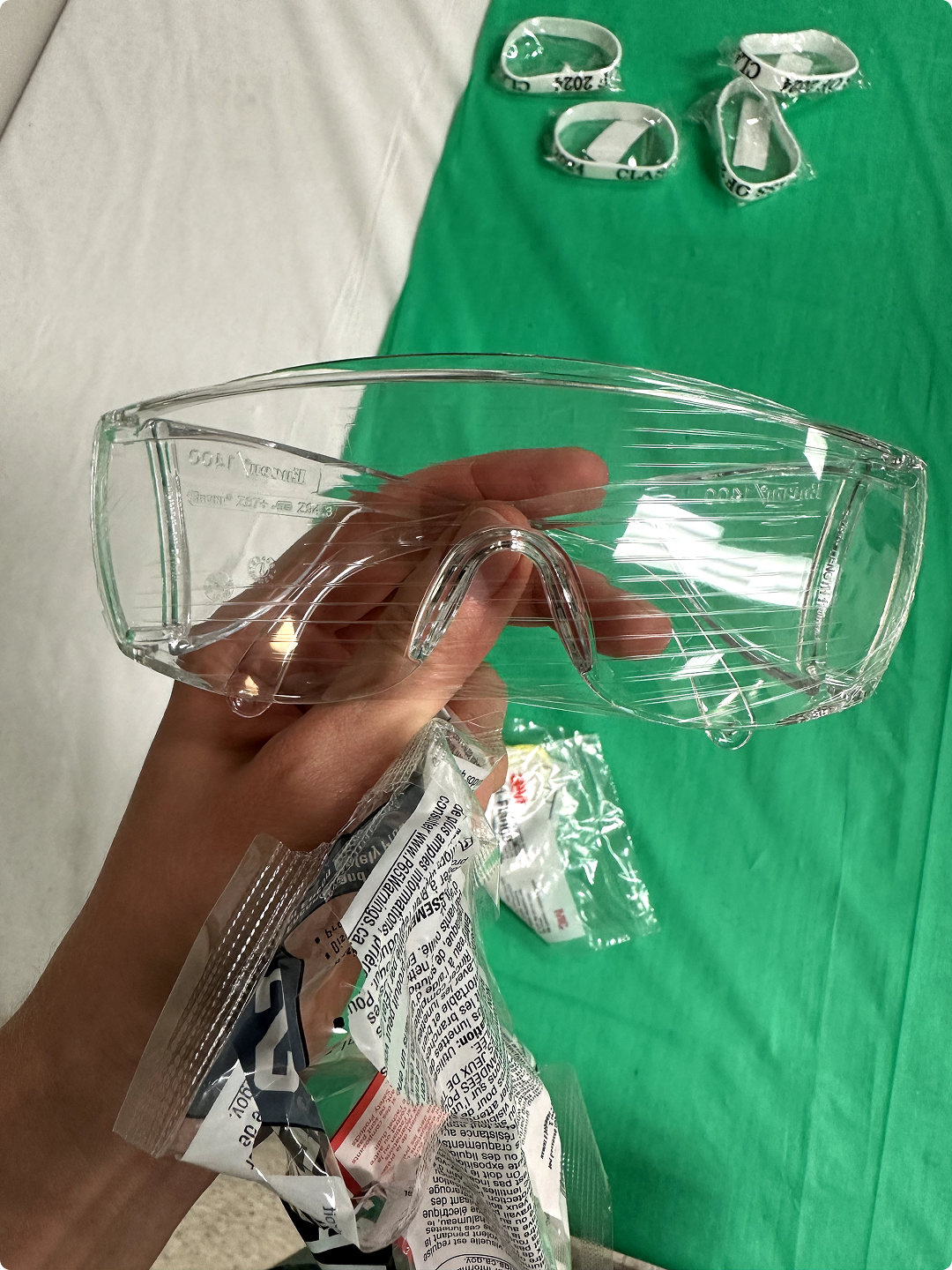 Hand holding clear safety goggles with wristbands on a green and white background.