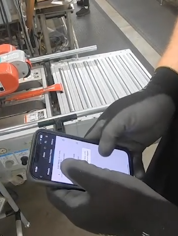 Person in black gloves holding smartphone over machinery in an industrial setting.