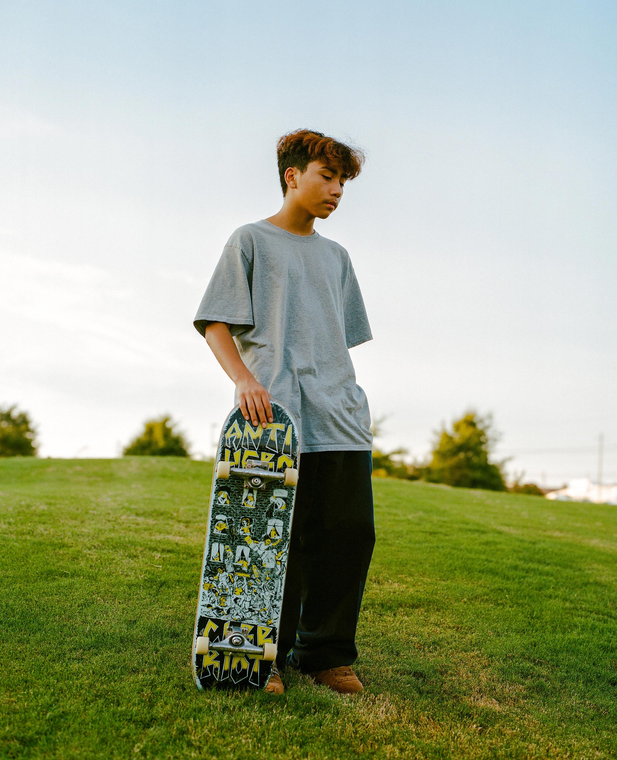 Teen boy in gray shirt holding a skateboard standing on green grass with blue sky background.