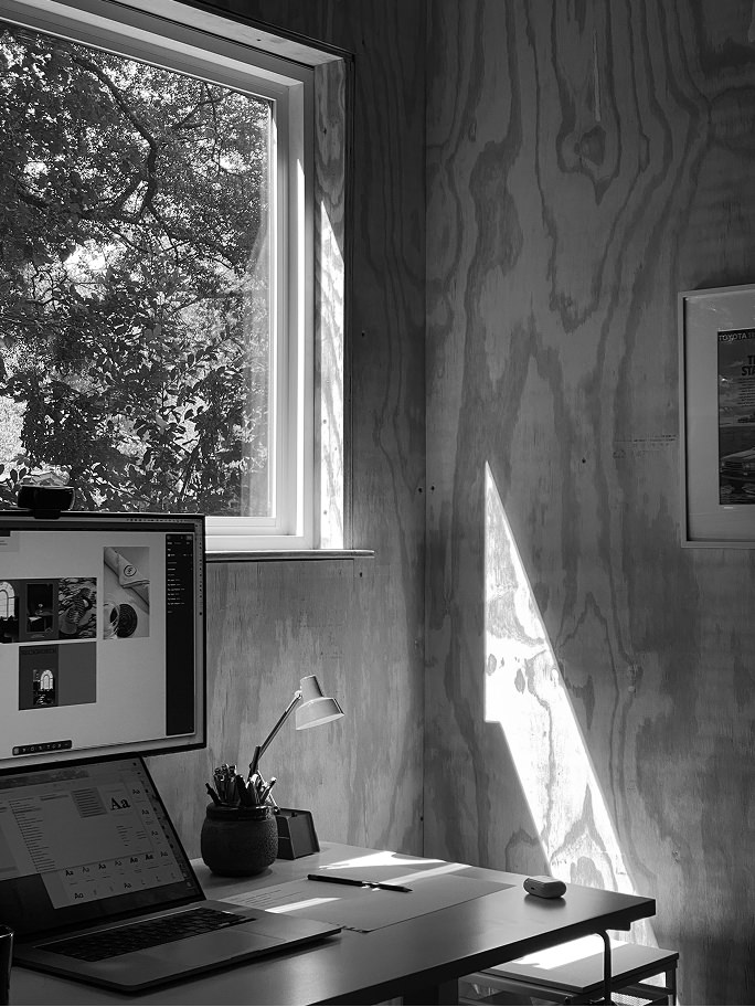 Sunlit workspace with a laptop, monitor, desk lamp, and window showing leafy trees outside.